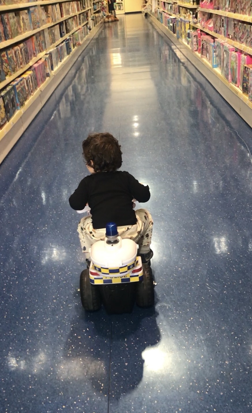 A young child with dark, curly hair seen from behind, riding a small white and blue toy police motorcycle down the long, shiny blue aisle of a toy store, exploring independently